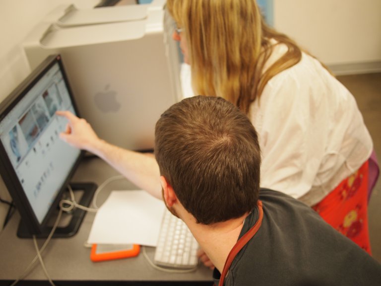 An artist seated in front of a computer with another co-artist, engaged in digital work that represents the creative and business process of producing and selling art online, photo by Crista Cloutier.